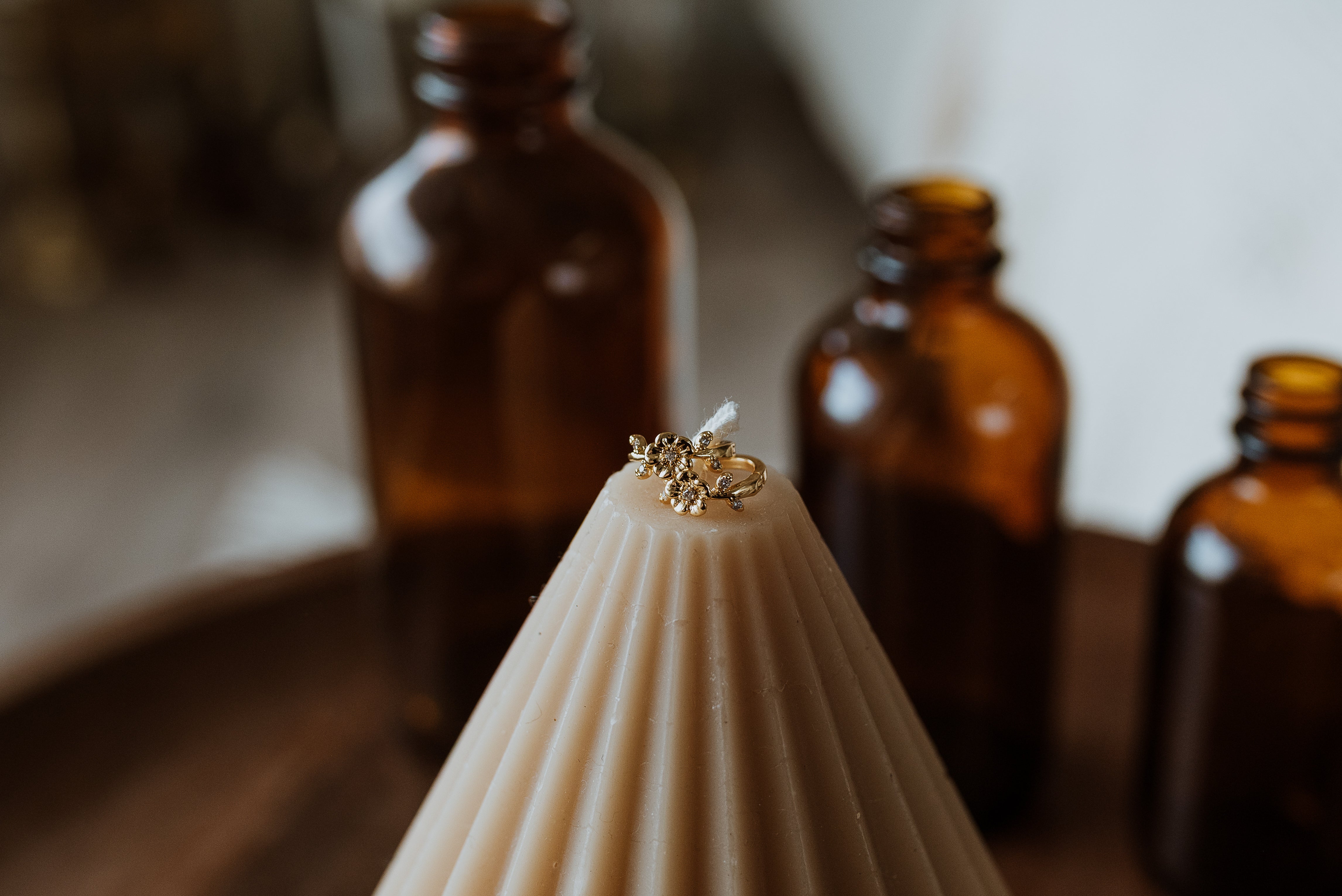 Candle with gold earrings on top in front of amber glass bottles.