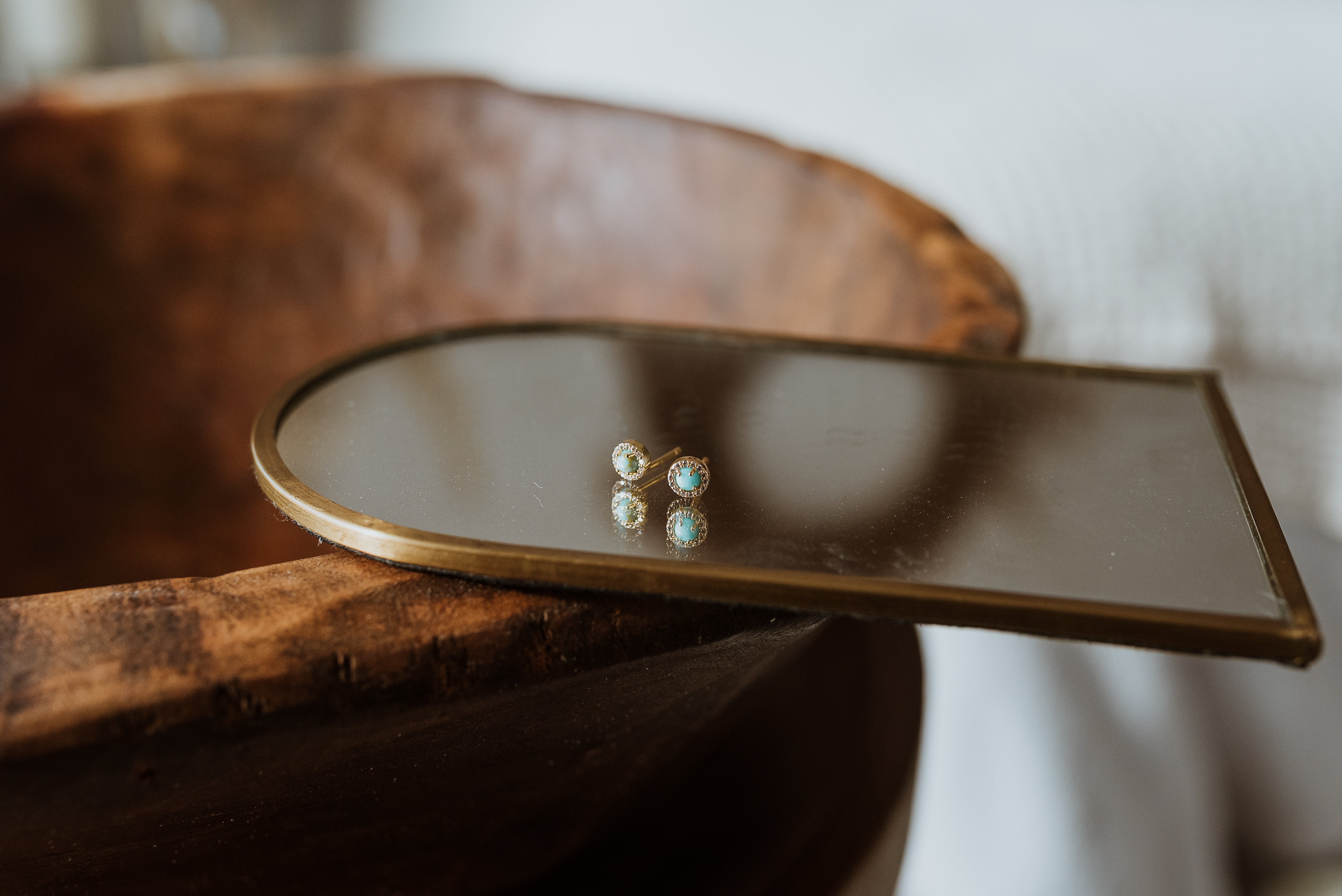 Small round mirror with gold frame on a wooden surface, reflecting turquoise earrings.