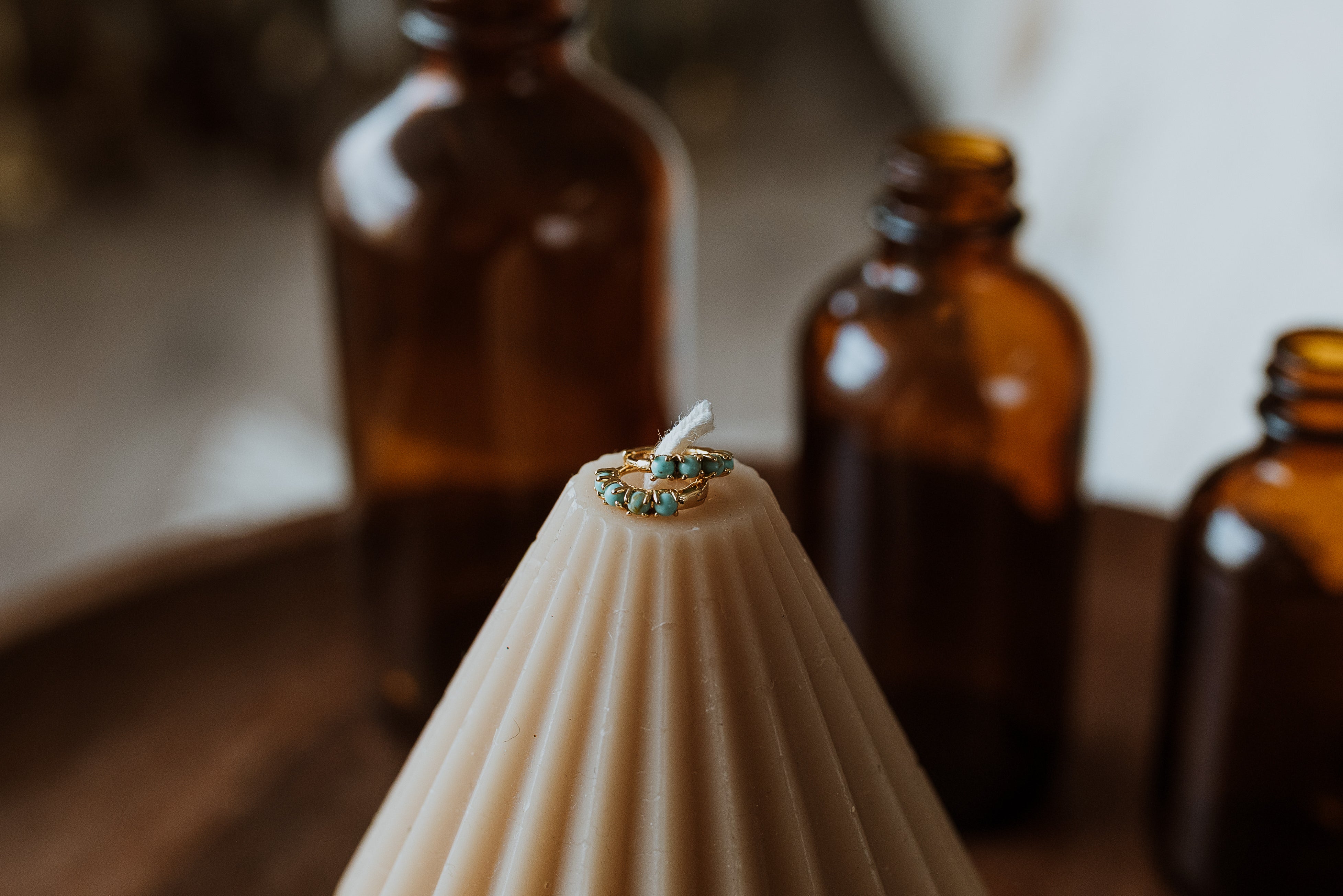 White candle with turquoise stone earrings on a wooden surface with brown bottles in the background