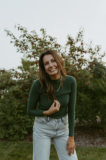Woman wearing a green sweater and light blue jeans standing outdoors with trees in the background while wearing gold jewellery.