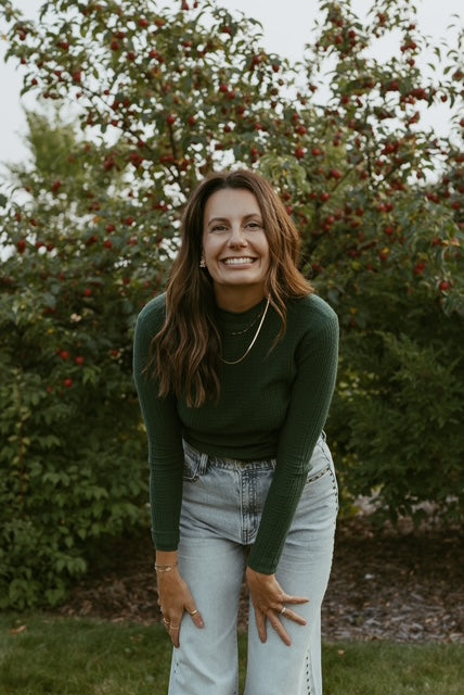 Woman in a green sweater and light jeans standing in front of trees with red berries while wearing gold jewellery.